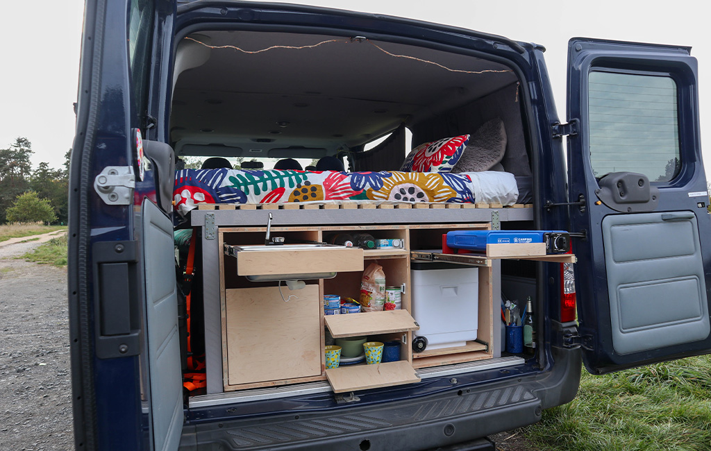 Geöffneter Van mit ausgezogener Campingküche, sichtbar sind Gasherd, Spüle und Vorratsfächer mit Lebensmitteln.