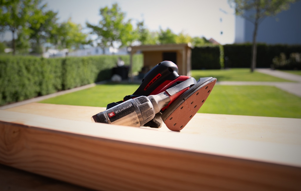 Ein Einhell Akku-Multischleifer steht auf einem Holzbalken im Freien auf einer Rasenfläche.
