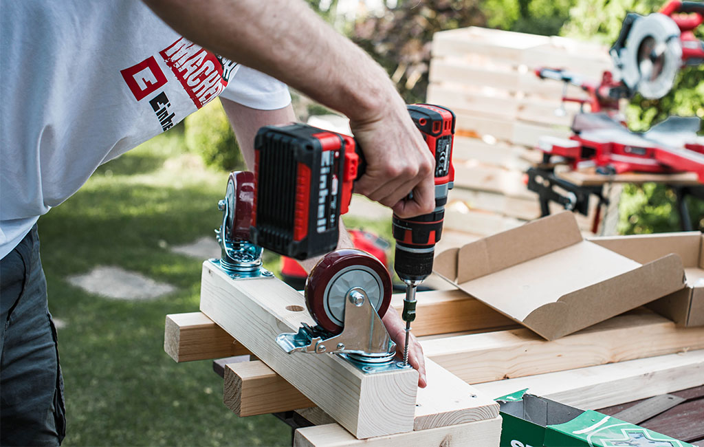 A man uses a cordless drill to attach large wheels to a wooden frame.