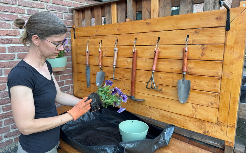 Eine Frau arbeitet an einem selbstgebauten Pflanztisch mit Rückwand, Haken und Gartenwerkzeugen beim Einpflanzen.