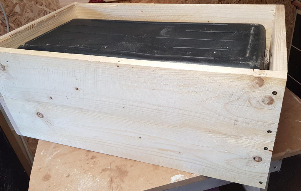 Eine schwarze Kunststoffbox ist in einen rechteckigen Holzkasten mit hellen Holzlatten eingesetzt.
