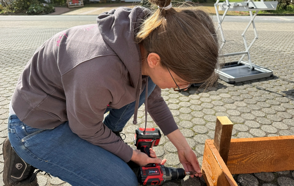 Eine Frau kniet auf Pflastersteinen und verschraubt zwei Holzbretter mit einem Einhell Akku-Bohrschrauber.