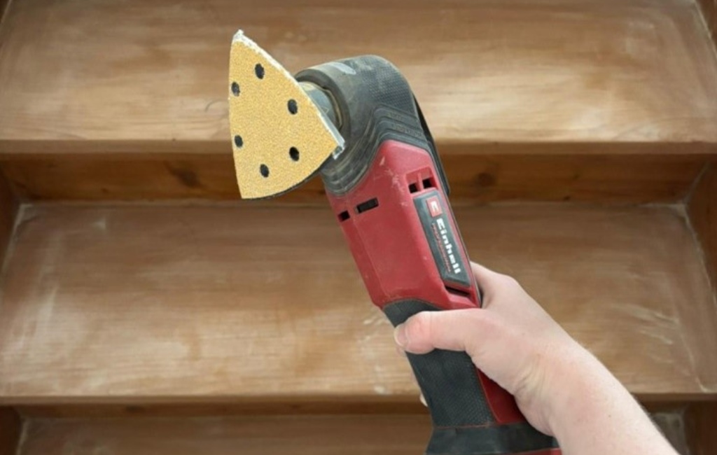Eine Hand hält ein rotes Einhell Multitool mit dreieckigem Schleifaufsatz vor einer Holzstufe.