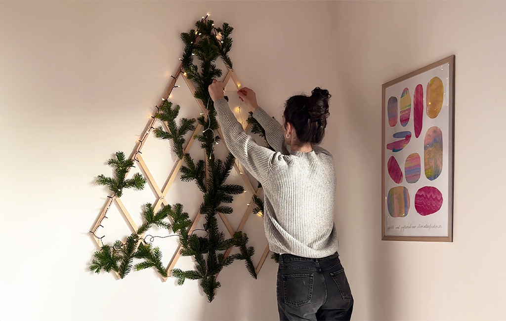 Eine Frau dekoriert ein Holzgitter in Tannenbaumform an der Wand mit Tannenzweigen und Lichterkette.