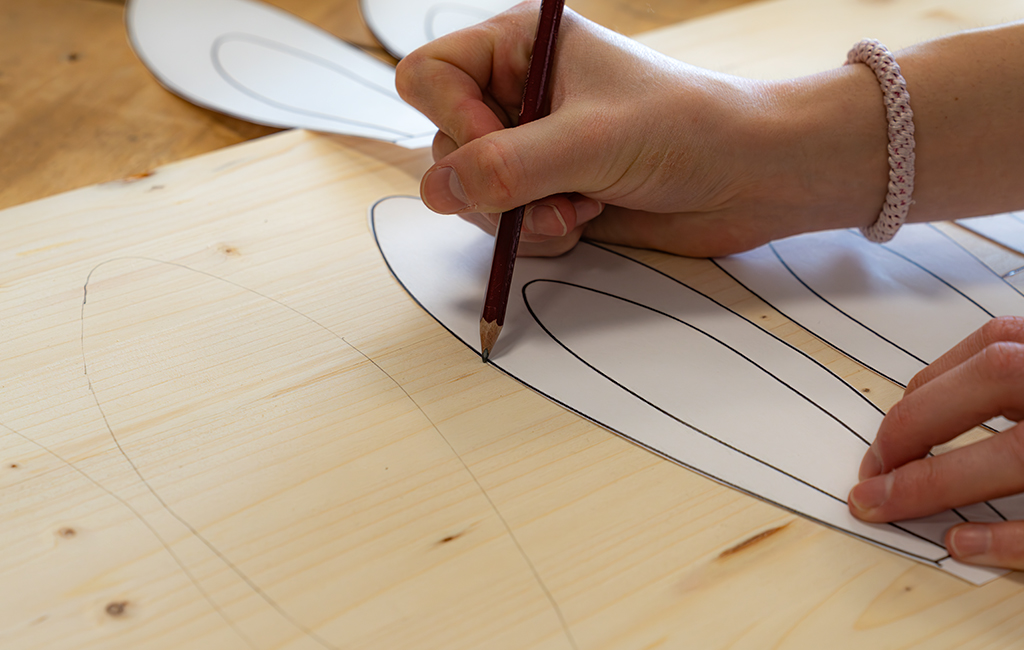 A hand traces rabbit ear outlines with a pencil onto a wooden board using a template.