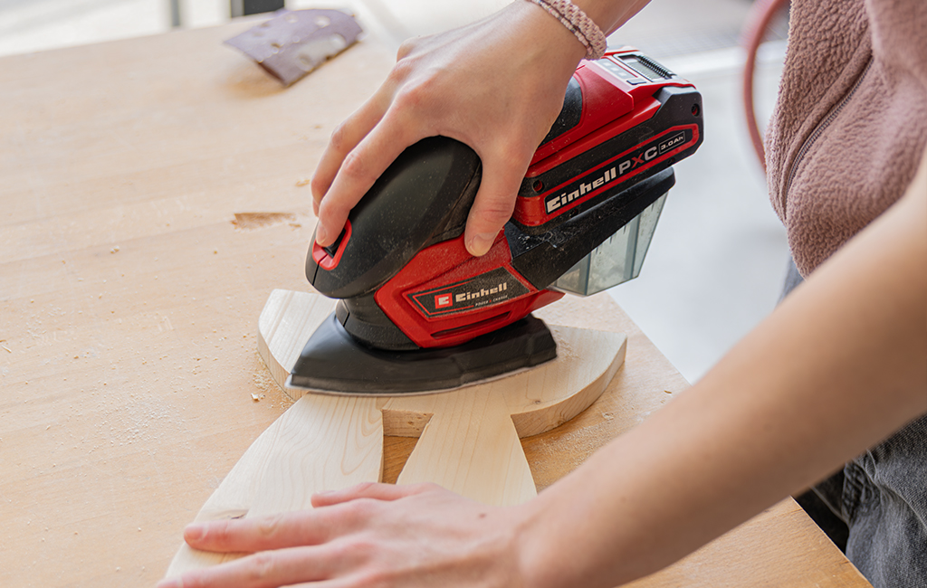 A person sands a cut wooden shape with an electric sander on a workbench.