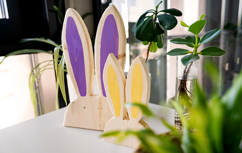 Two decorative wooden rabbits with painted ears stand on a table next to indoor plants.