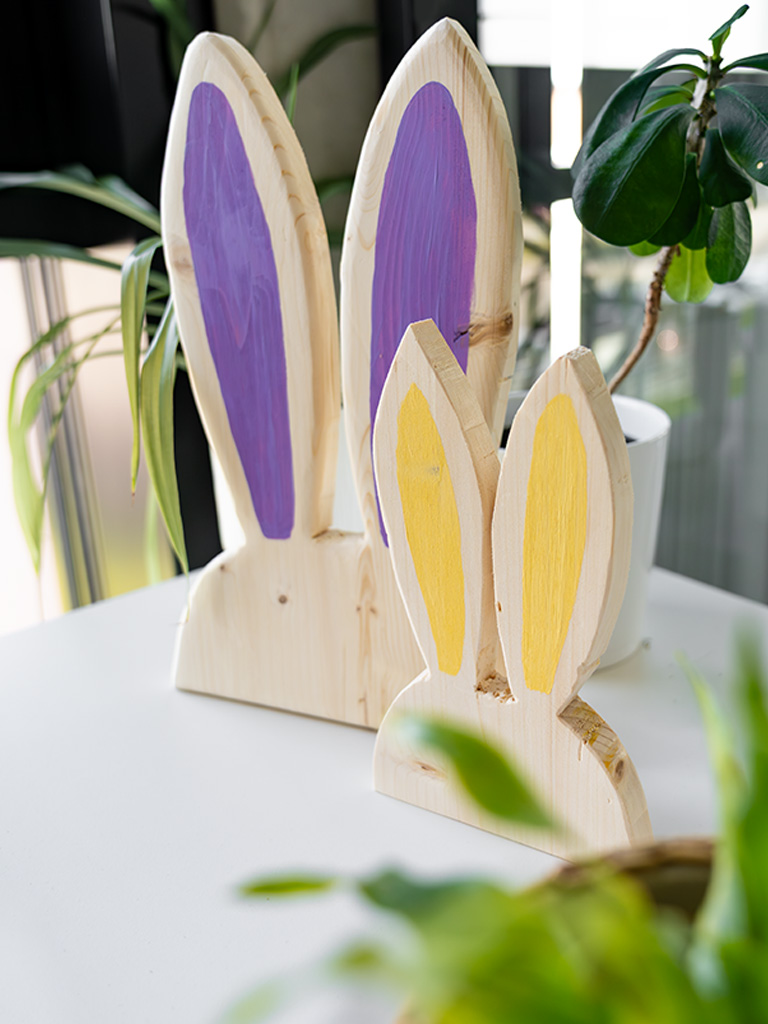 Two decorative wooden rabbits with painted ears stand on a table next to indoor plants.
