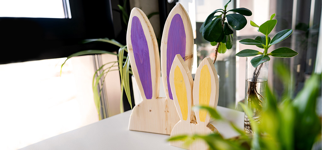Two decorative wooden rabbits with painted ears stand on a table next to indoor plants.