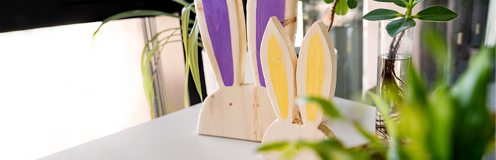 Two decorative wooden rabbits with painted ears stand on a table next to indoor plants.