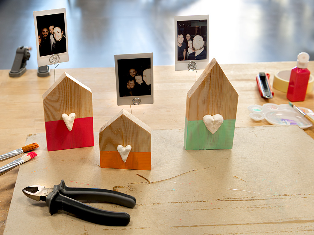 Three house-shaped wooden photo holders with colored bases and white hearts, arranged on a craft table.