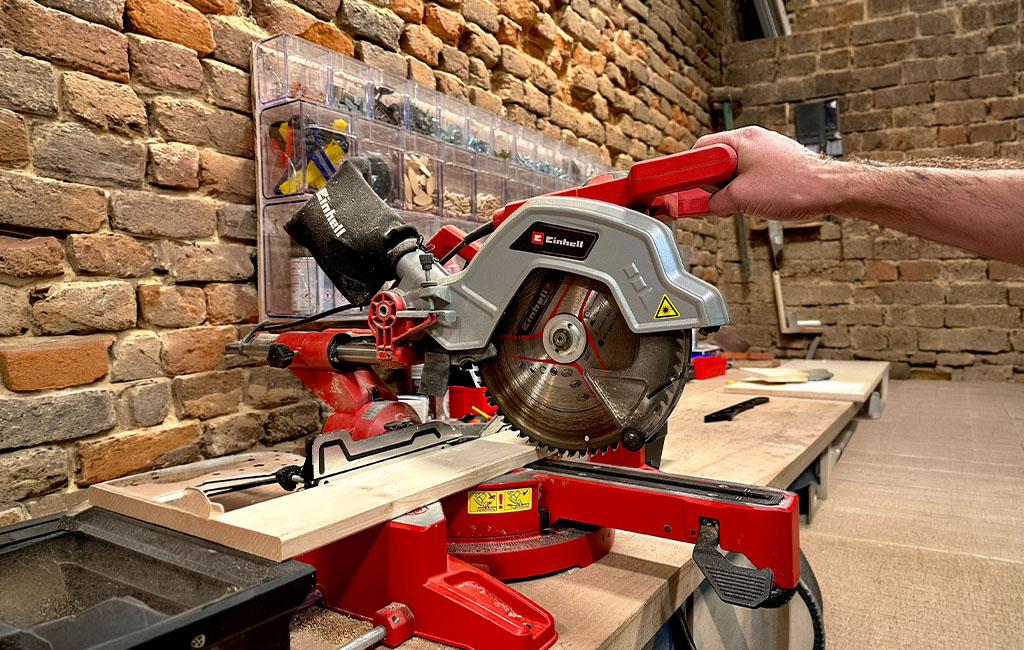 Mitre saw cutting a wooden board on a workbench in a workshop, with the circular saw blade lowered onto the wood during cutting.