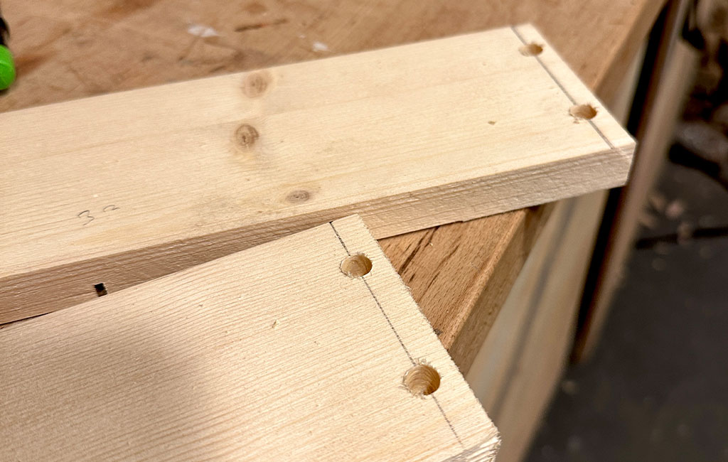 Two wooden boards with drilled holes and marked lines lying on a workbench, prepared for assembly.