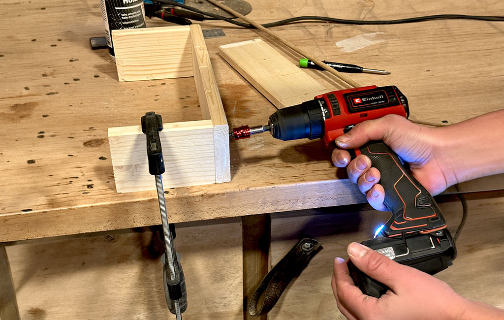 Person fastening wooden parts with a cordless screwdriver on a workbench, with the boards fixed in place using a clamp.