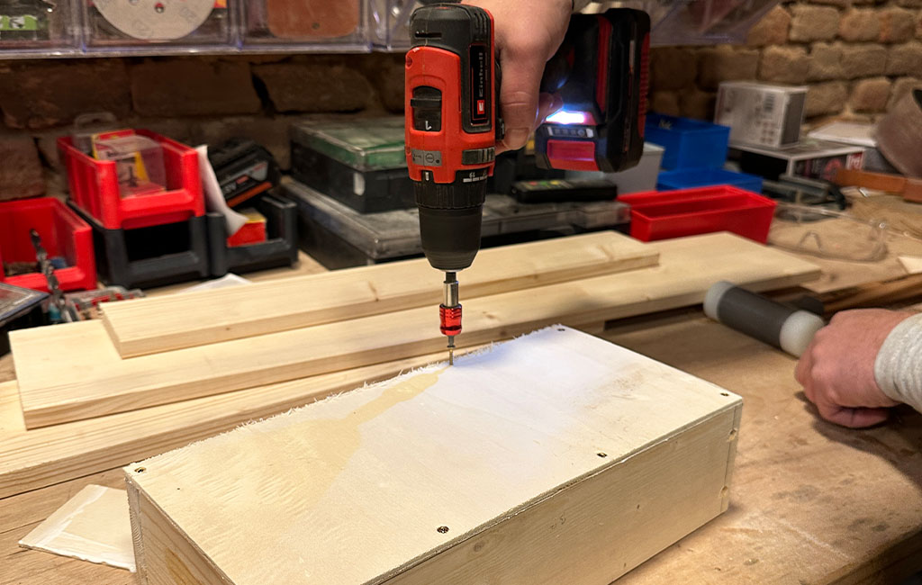 Cordless screwdriver fastening screws into the lid of a wooden box on a workbench while the box is being assembled.
