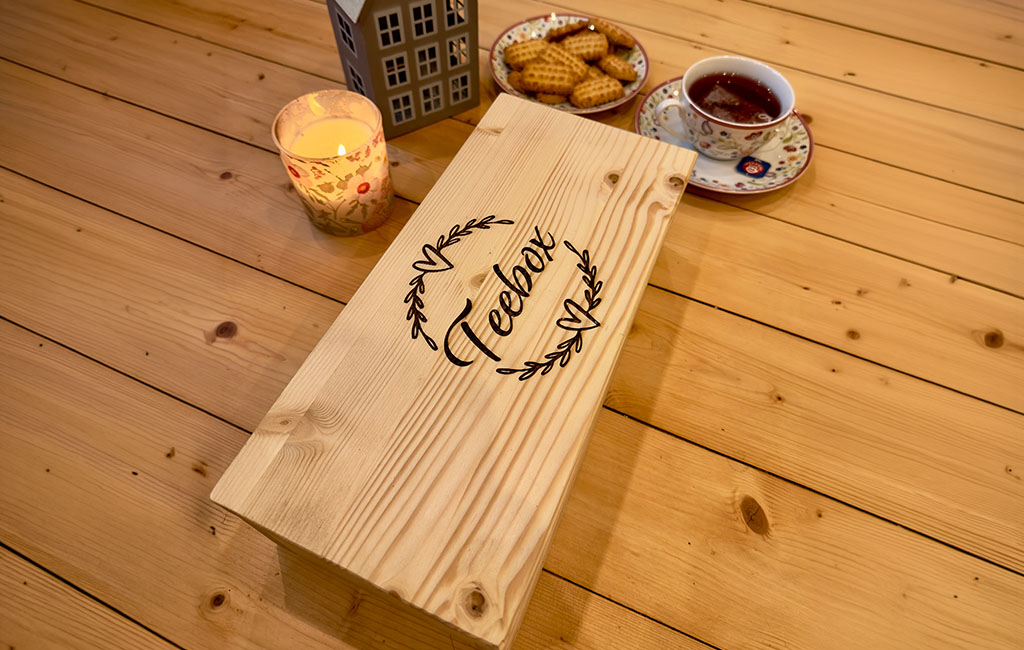Wooden tea box with engraved “Teebox” on the lid placed on a table, next to a cup of tea, cookies and a lit candle.