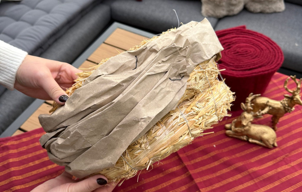 A straw wreath wrapped in brown paper is held over a table with Christmas decorations.