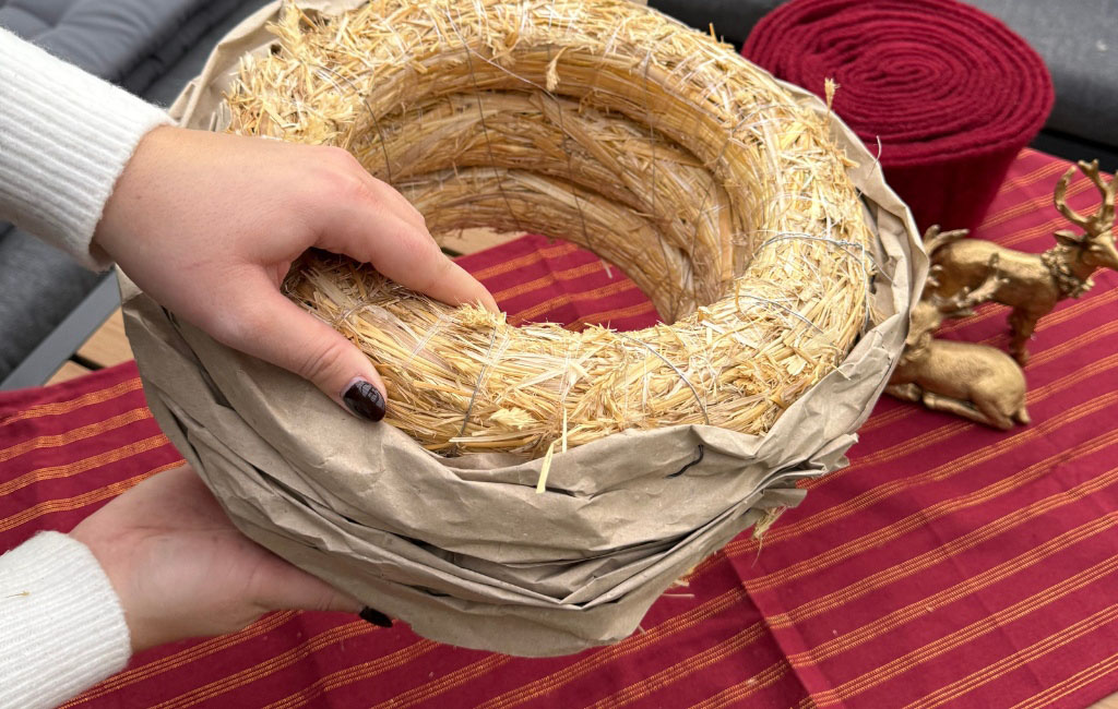 A paper-covered straw wreath is held by two hands, with red felt visible in the background.