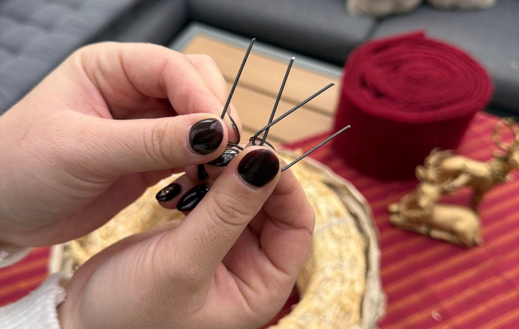 Two hands hold several silver wire clips for crafting, with a straw wreath in the background.