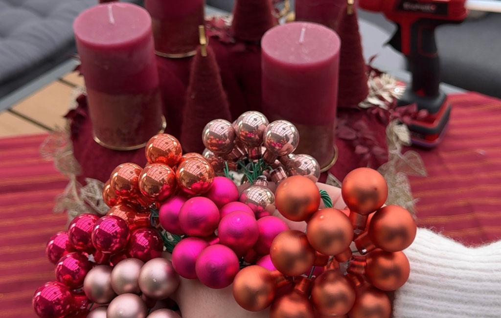 A hand holds red and pink mini Christmas baubles, with the Advent wreath visible in the background.