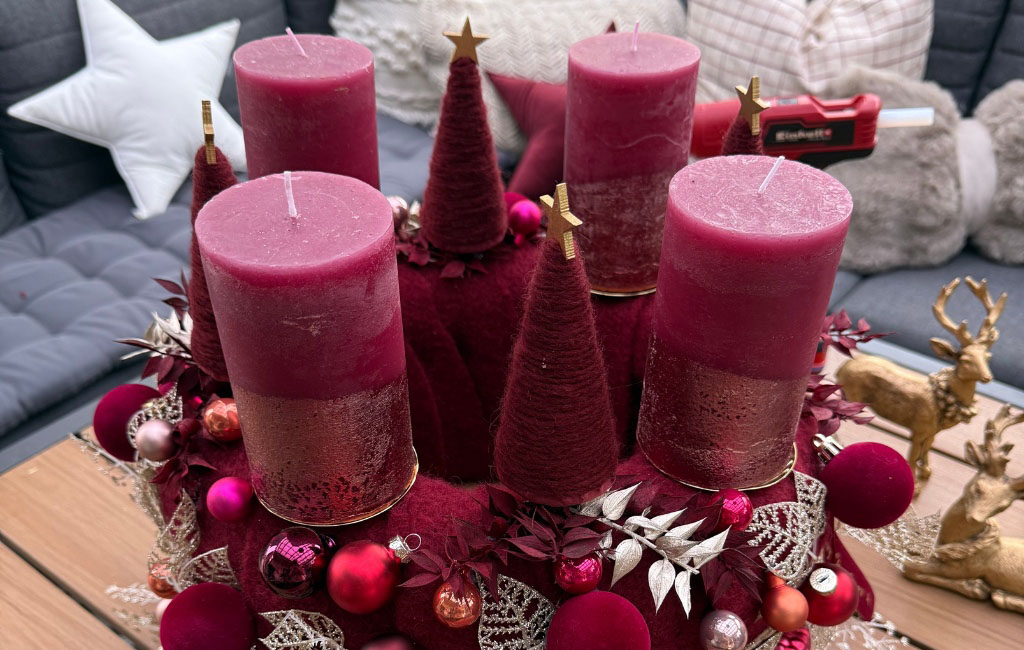 Der fertige Adventskranz mit vier roten Kerzen, Filzbäumen und Weihnachtskugeln steht dekorativ auf einem Tisch.