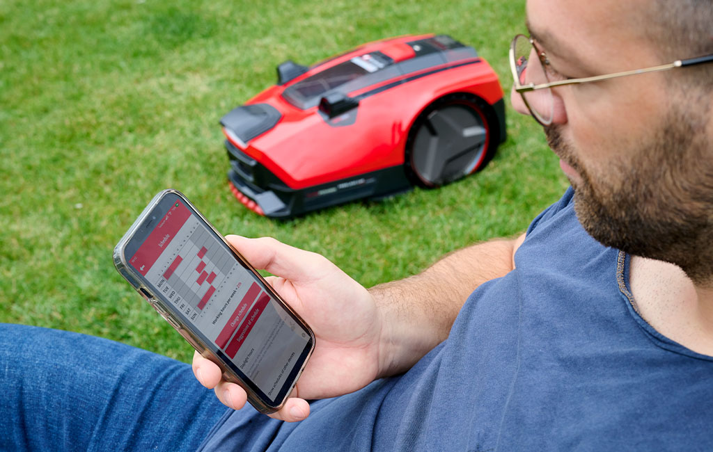 A man sits on the lawn and controls the Einhell robotic lawn mower FREELEXO CAM with an app on his smartphone.