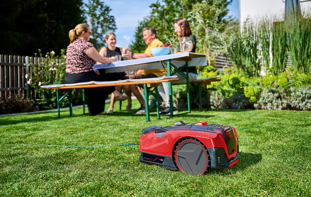 The Einhell robotic lawn mower FREELEXO CAM mows the lawn while four people sit at a garden table in the background.