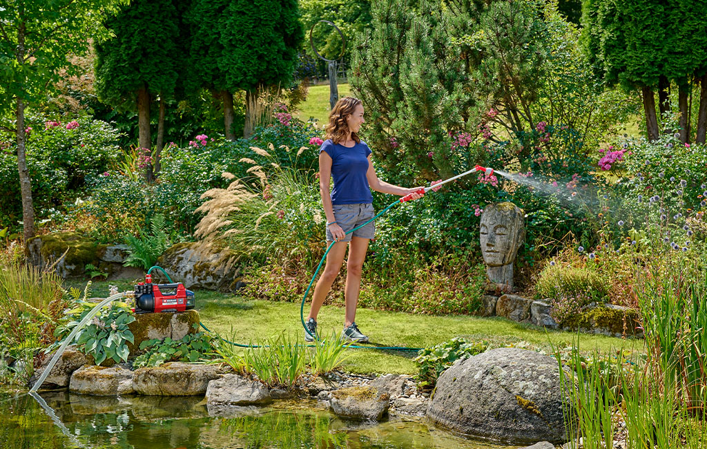A woman waters plants with a spray lance at a garden pond, with a Einhell garden pump beside her.