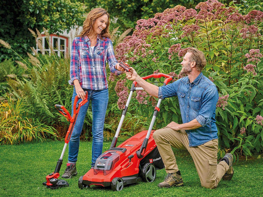 A woman with an Einhell cordless lawn trimmer handing a battery suitable for the cordless lawn mower to the man next to her.