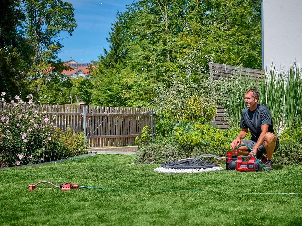 Ein Mann kniet neben einer schnurlosen Gartenpumpe, die Wasser zu einem Rasensprenger pumpt. 