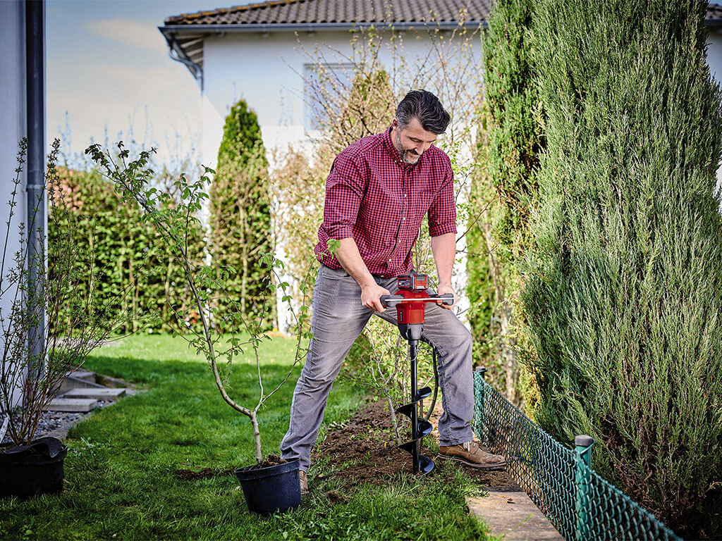 Ein Mann bohrt mit dem Akku-Erdbohrer von Einhell ein Loch in den Gartenboden.