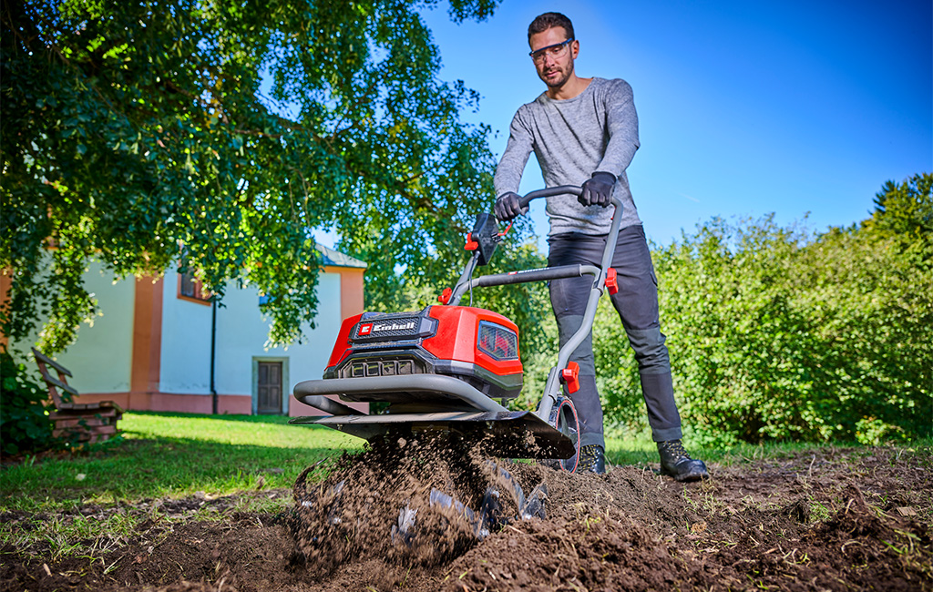 Man wearing safety glasses operates a red tiller, loosening soil in a garden in front of a house.