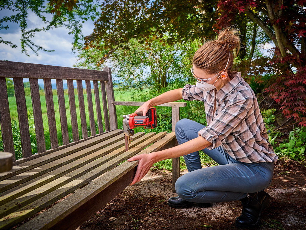 Frau im Garten beim Schleifen von engen Zwischenräumen einer Holz-Gartenbank mit einer Akku-Bandfeile