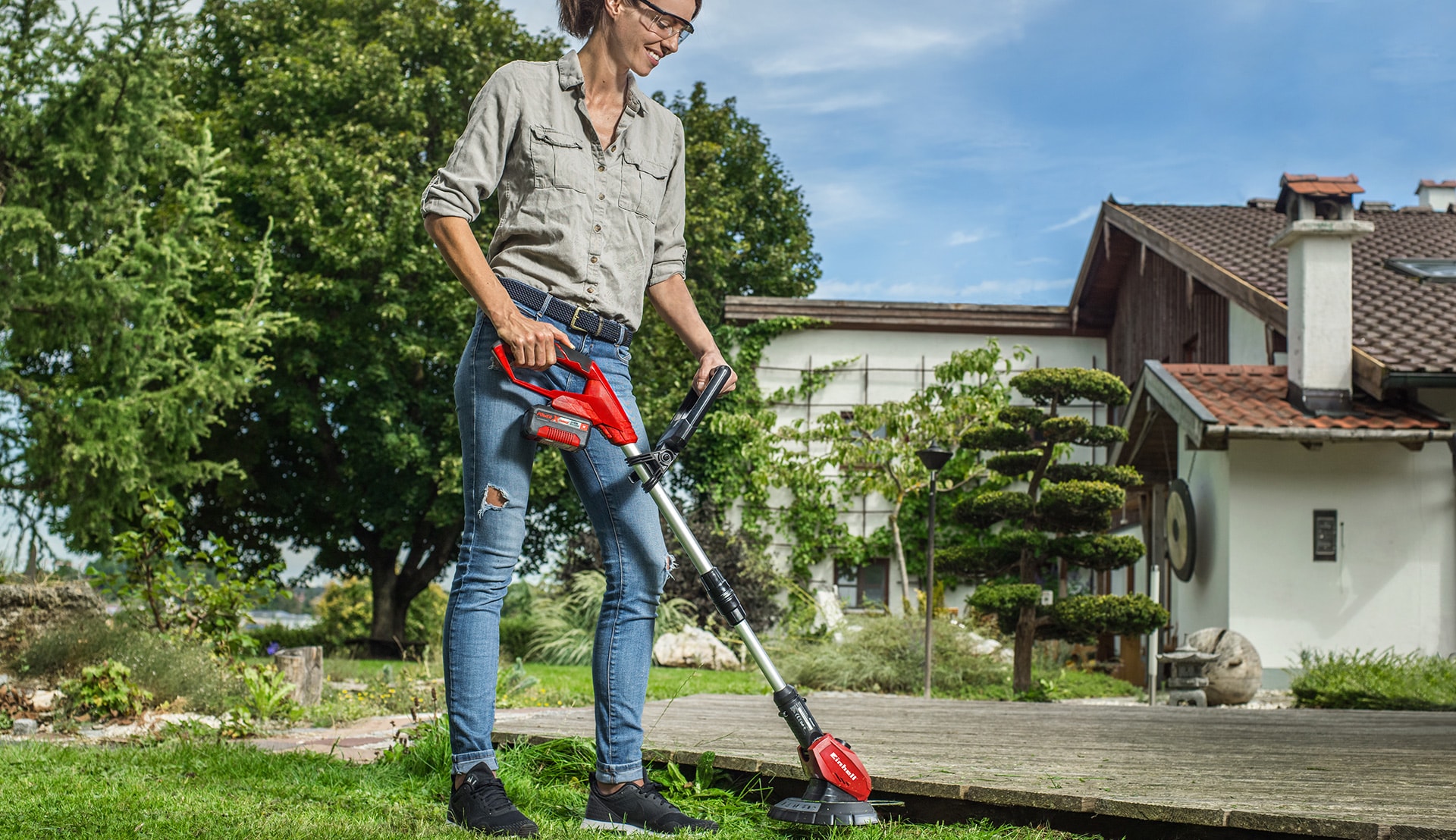 A woman trims the grass under a footbridge with an Einhell grass trimmer.