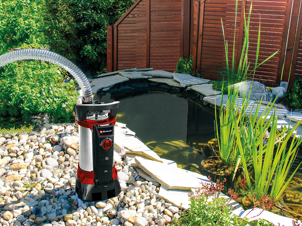 Eine Einhell-Schmutzwasserpumpe steht neben einem Gartenteich mit Steinen, Pflanzen und kleinem Wasserfall.all waterfall.