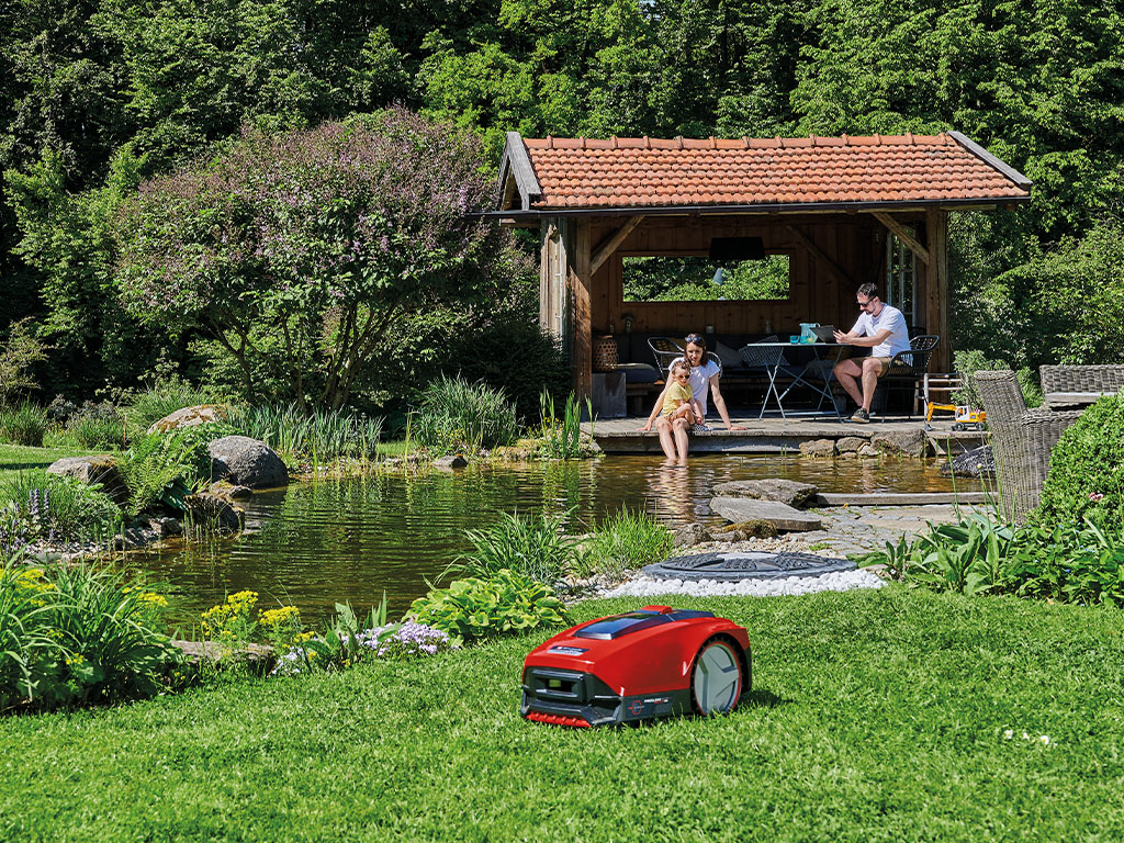 A robotic mower cuts grass in a large garden with a covered terrace and a pond in the background.