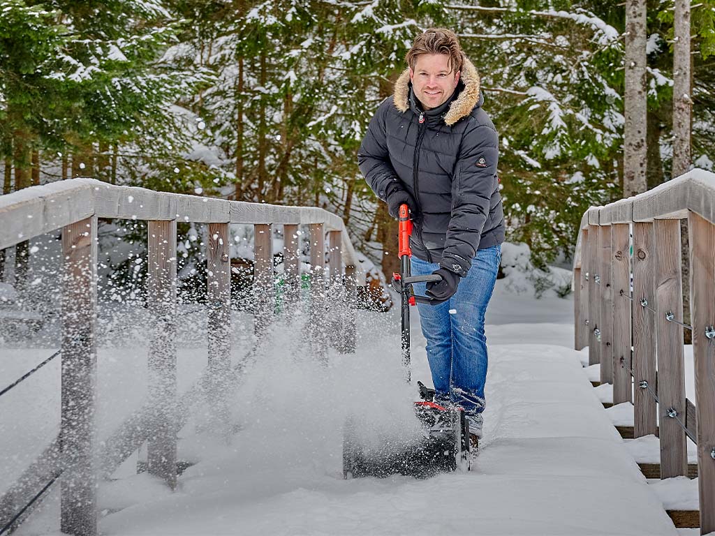 Mann beim Schneeräumen mit einer Akku-Schneefräse auf einer schneebedeckten Brücke vor einem Wald