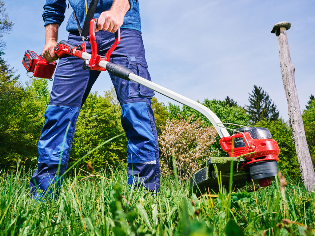 A man with blue trousers and a red cordless lawn trimmer trimming the grass in a meadow.