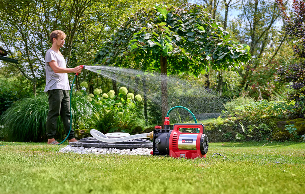 Ein Mann bewässert barfuß den Garten mit einer Einhell Gartenpumpe, die auf dem Rasen steht.