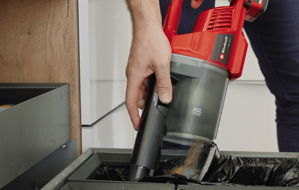 Hand empties the dust container of a red handheld vacuum with “Einhell” logo over a bin; “MAX” marking visible.