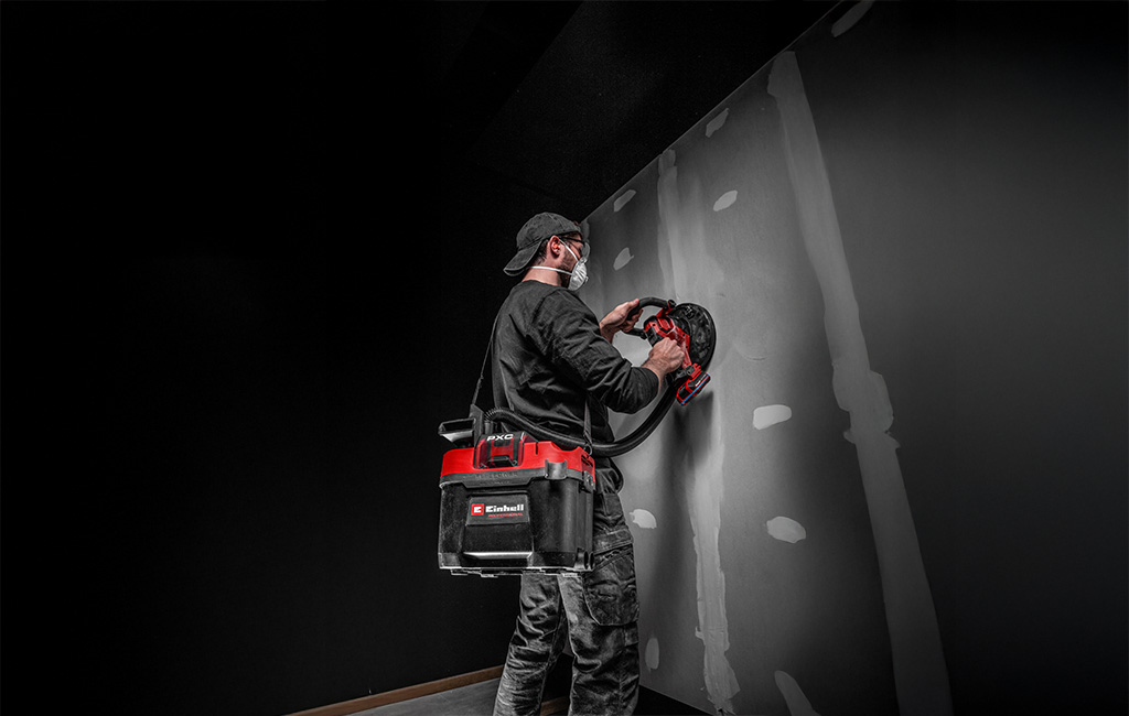 Eine Person saugt mit einem kabellosen Staubsauger eine Wand entlang, Staubpartikel werden sichtbar eingesogen.