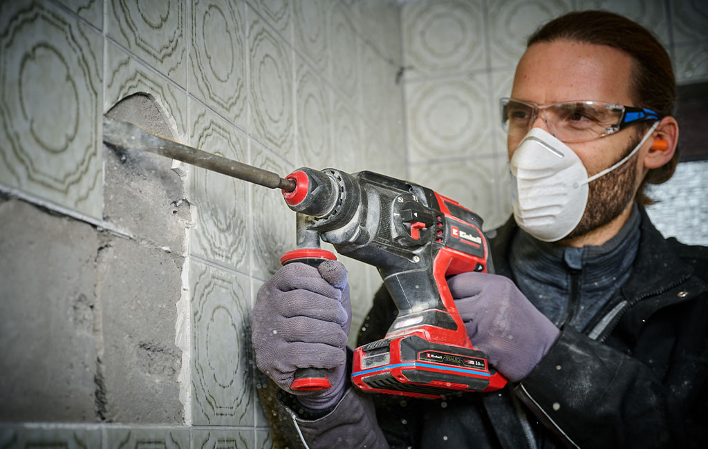 Ein Handwerker mit Schutzbrille und Atemmaske arbeitet mit einem Einhell Akku-Bohrhammer an einer gefliesten Wand.