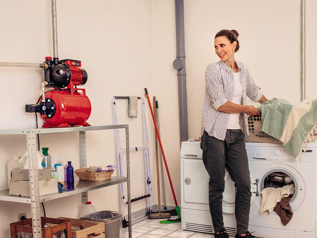 A woman stands in front of the washing machine with her laundry basket and looks at the connected Einhell domestic waterworks.