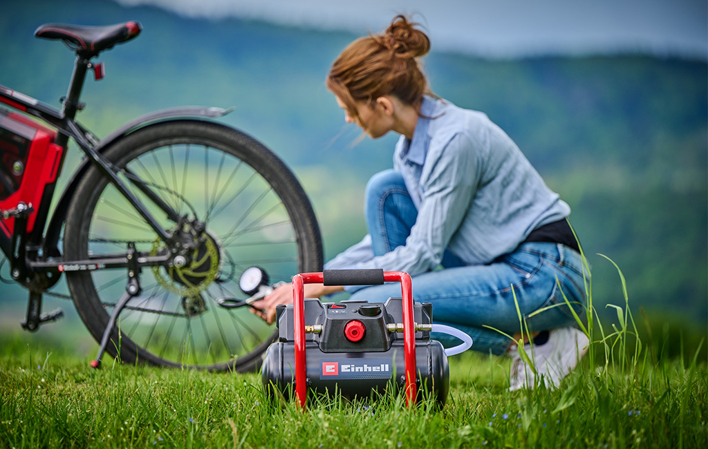 A woman inflates a bicycle tire with a cordless Einhell compressor while sitting in a green field.