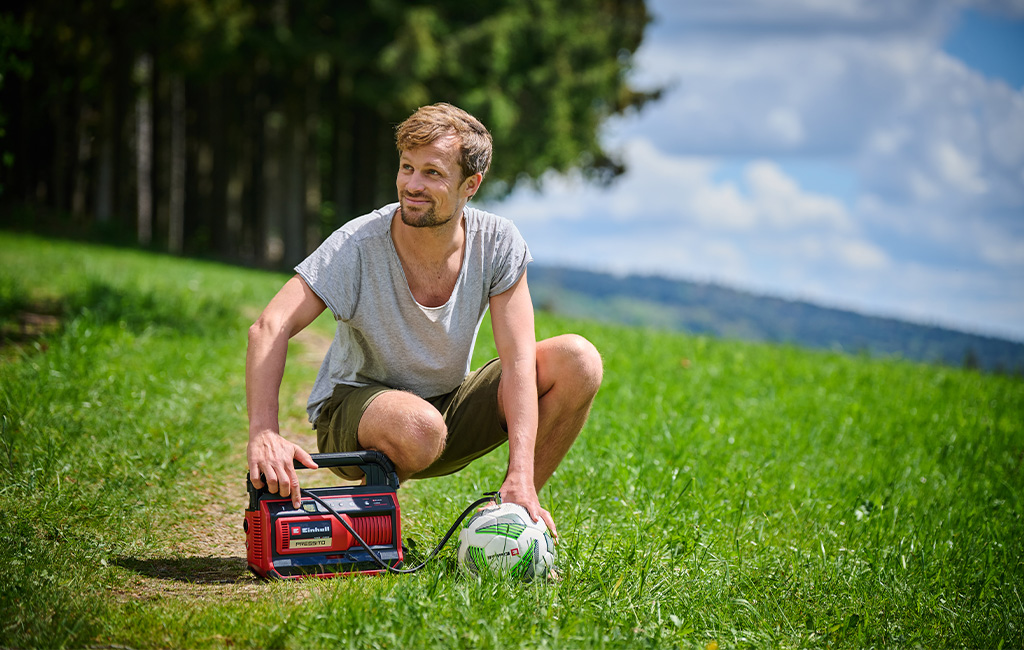 A man kneels on a lawn and uses the cordless Einhell compressor PRESSITO to inflate a soccer ball.