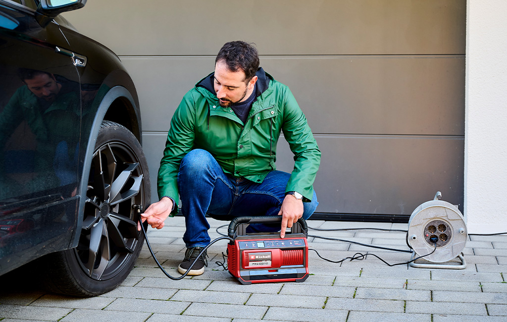 A man inflates a car tire with a compressor while kneeling in front of a garage.