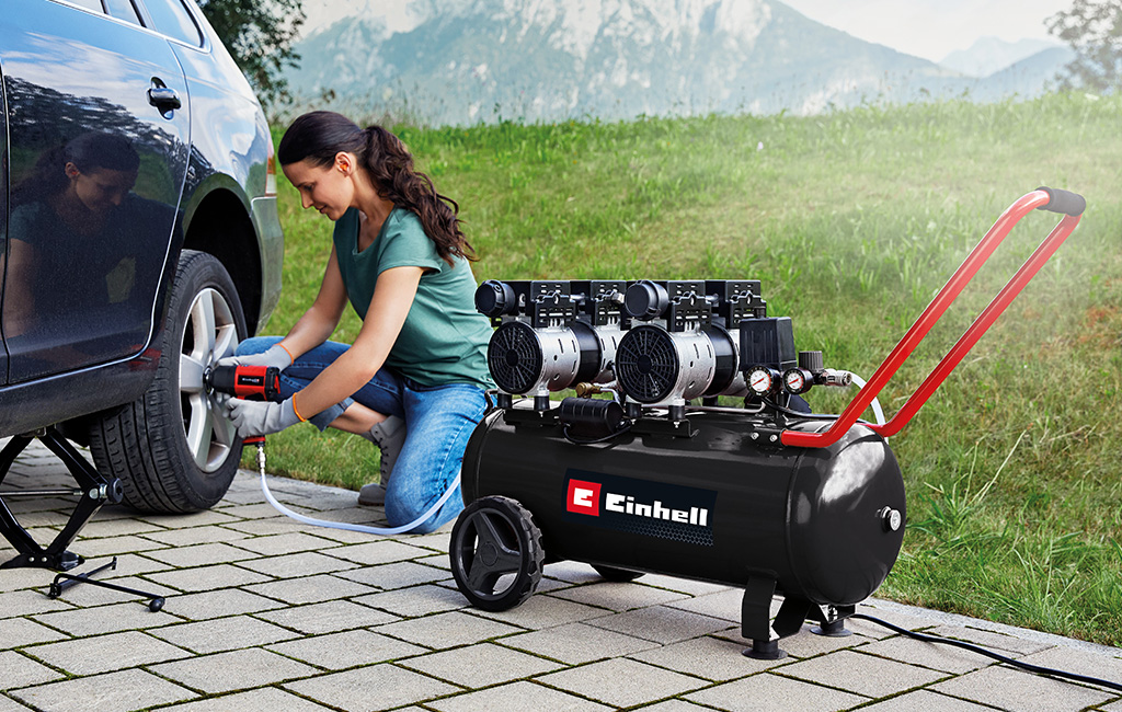 A woman uses a Einhell compressor to inflate car tires while sitting on a driveway.