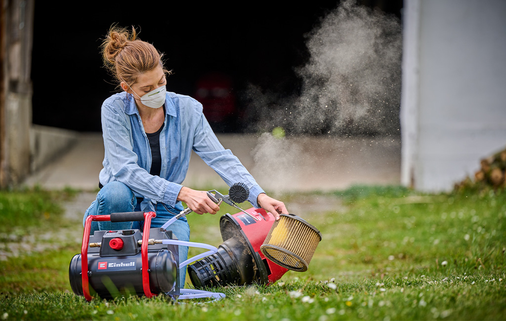 A woman cleans a dusty filter with a Einhell portable compressor outdoors.