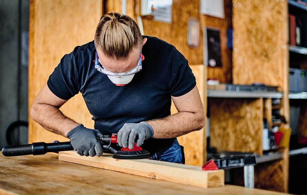 A man sands a wooden board using a pneumatic sanding tool in a workshop.