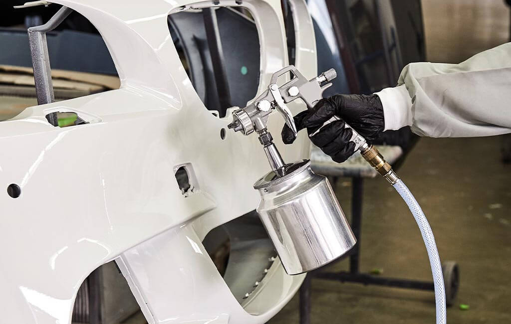 Close-up of a person using a spray gun to paint a bumper of a car.